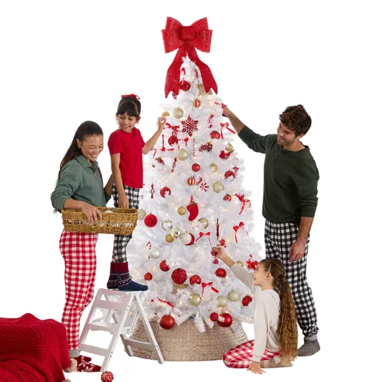 A family decorates a white Christmas tree with red and gold ornaments in a cozy living room setting. They wear plaid pajamas, and the room features a fireplace and holiday decorations.