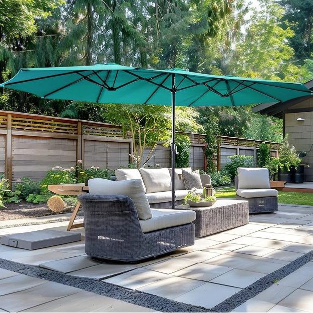 Outdoor patio set featuring a wicker-style sofa and chairs with cushions, a rectangular coffee table, and a large green patio umbrella. Surrounded by a fenced garden with greenery and a small house in the background.