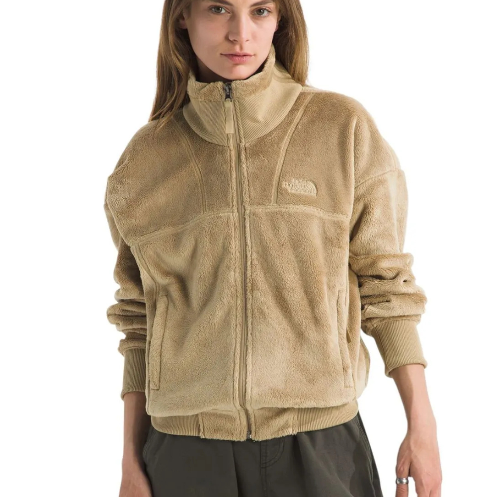 A woman wears a tan, fuzzy fleece jacket featuring a front zipper and The North Face logo embroidered on the chest, paired with dark pants.