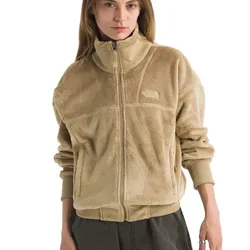A woman wears a tan, fuzzy fleece jacket featuring a front zipper and The North Face logo embroidered on the chest, paired with dark pants.