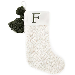 A white knit Christmas stocking with a textured pattern, featuring a green letter \“F\“ on the cuff, adorned with green pom-poms and tassels.