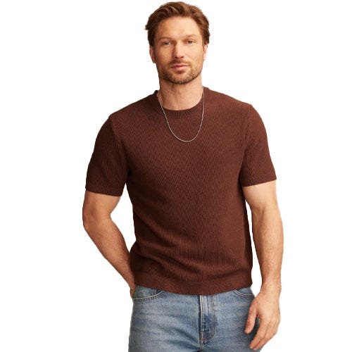 A man is wearing a short-sleeve brown ribbed sweater and light blue jeans, accessorized with a silver necklace.