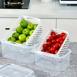 Two white perforated storage bins with green apples and red tomatoes inside are placed on a kitchen counter, accompanied by their transparent lids.