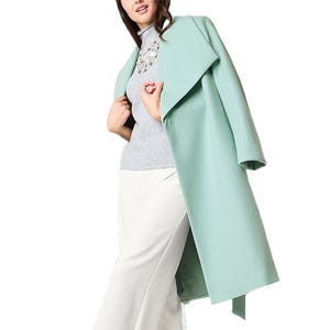 A woman models a stylish outfit featuring a light teal overcoat, a gray high-neck top with lace detailing, and white pants.