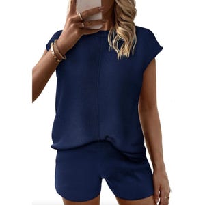 A woman is wearing a navy blue short-sleeve top and matching shorts, accessorized with bracelets and a ring, while holding a phone.