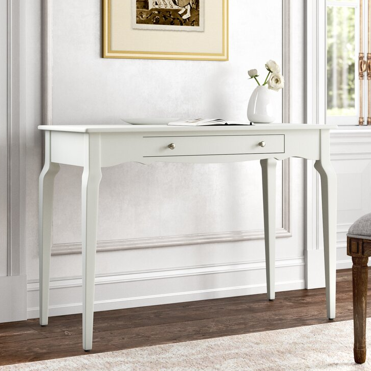 A white writing desk with elegant curved legs, a central drawer featuring two small knobs, and a flat top surface. It is positioned against a classic wall, with a framed artwork above and a white vase with flowers on top.