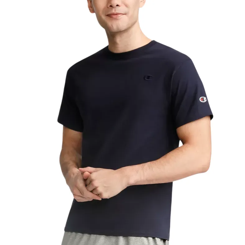 A person wears a navy blue Champion t-shirt with the brand's logo on the chest and sleeve, paired with gray pants.