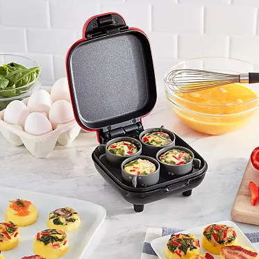 A countertop appliance is shown cooking mini frittatas, with ingredients like eggs and vegetables surrounding it, including a bowl of whisked eggs, spinach, and red pepper slices.