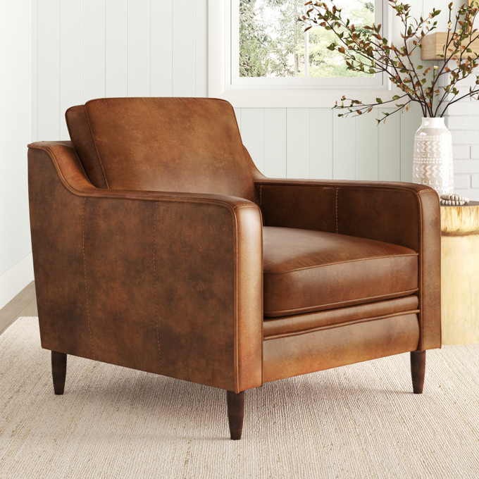 A brown, genuine leather deep-seat chair with a smooth finish features angled arms and tapered wooden legs, exuding a classic and elegant style. It's set in a minimalistic room with light walls and natural decor.