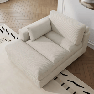 A cream-colored, corduroy-upholstered chaise lounge with a padded backrest and side armrest, featuring a rectangular throw pillow. The lounge sits on a wooden herringbone floor next to a patterned rug.