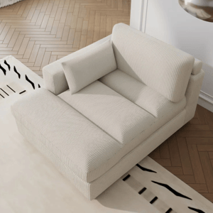 A cream-colored, corduroy-upholstered chaise lounge with a padded backrest and side armrest, featuring a rectangular throw pillow. The lounge sits on a wooden herringbone floor next to a patterned rug.