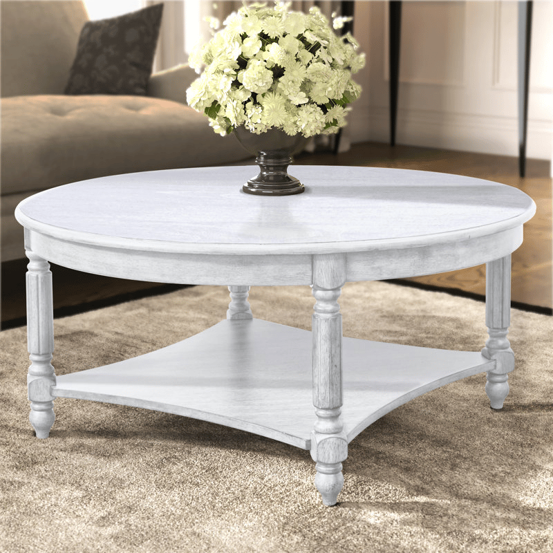 Round, solid wood coffee table with a distressed white finish, featuring turned legs and a lower shelf. A bouquet of white flowers in a vase is placed on top.