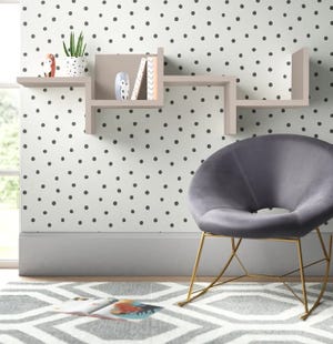 A modern, gray velvet accent chair with gold legs sits in front of a wall-mounted, asymmetrical bookshelf holding decorative items, against a polka dot wallpaper. A geometric patterned rug lies beneath them.