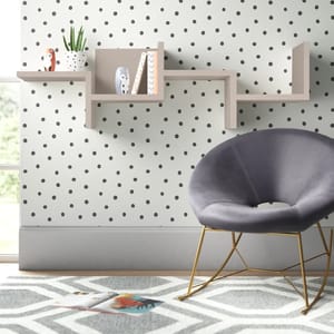 A modern, gray velvet accent chair with gold legs sits in front of a wall-mounted, asymmetrical bookshelf holding decorative items, against a polka dot wallpaper. A geometric patterned rug lies beneath them.