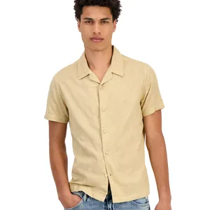 A man is wearing a beige short-sleeve button-up shirt with a subtle pattern, paired with blue jeans.