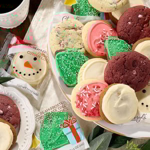 A variety of Cheryl's holiday cookies, including frosted tree-shaped cookies with green icing and sprinkles, red and white frosted round cookies with sprinkles, and a snowman face cookie, arranged on a plate.