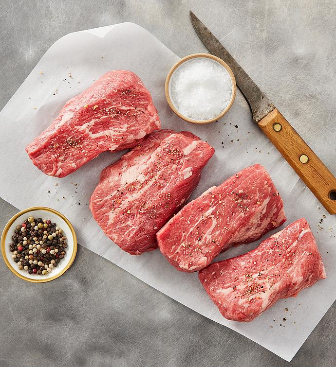Four grass-fed beef tri-tip steaks are on parchment paper, seasoned with salt and mixed peppercorns. A bowl of coarse salt and a wooden-handled knife are also present.