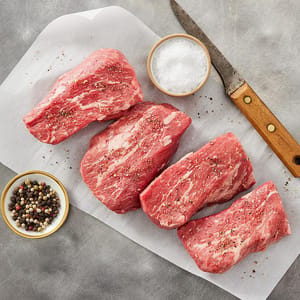Four grass-fed beef tri-tip steaks are on parchment paper, seasoned with salt and mixed peppercorns. A bowl of coarse salt and a wooden-handled knife are also present.