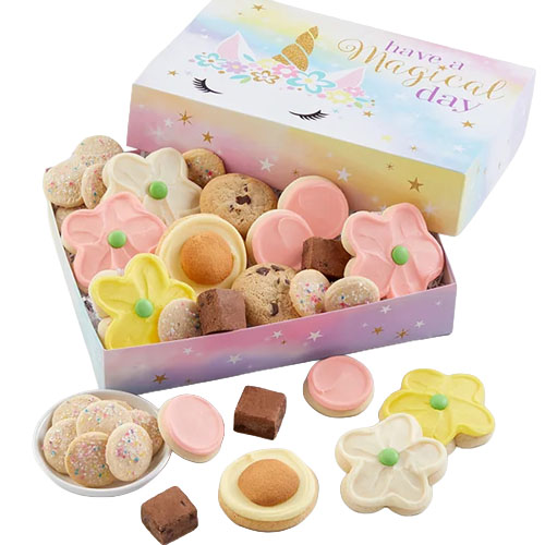 A pastel-colored box with \“Have a Magical Day\“ contains a variety of cookies, including flower-shaped, round pink, chocolate chip, and sprinkle-topped cookies, along with small fudge squares.