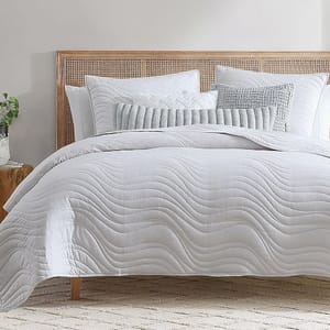 A neatly made bed with a cane headboard features a white, wavy-patterned comforter, white and light gray pillows, and a decorative throw pillow. A small plant sits on a wooden bedside table.