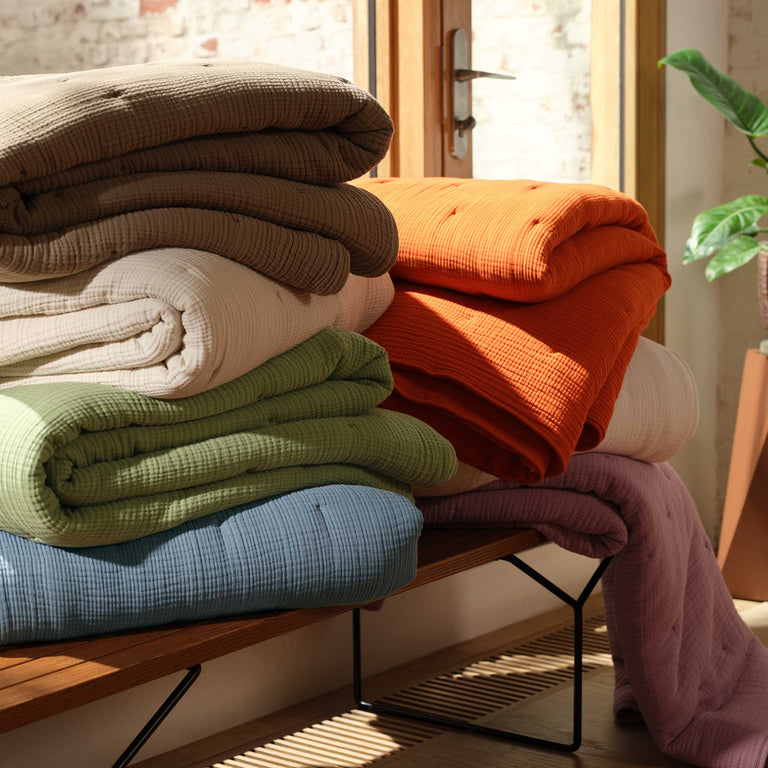 A stack of folded, textured blankets in various colors, including brown, orange, white, green, blue, and lavender, sits on a wooden bench in a sunlit room.