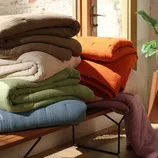 A stack of folded, textured blankets in various colors, including brown, orange, white, green, blue, and lavender, sits on a wooden bench in a sunlit room.