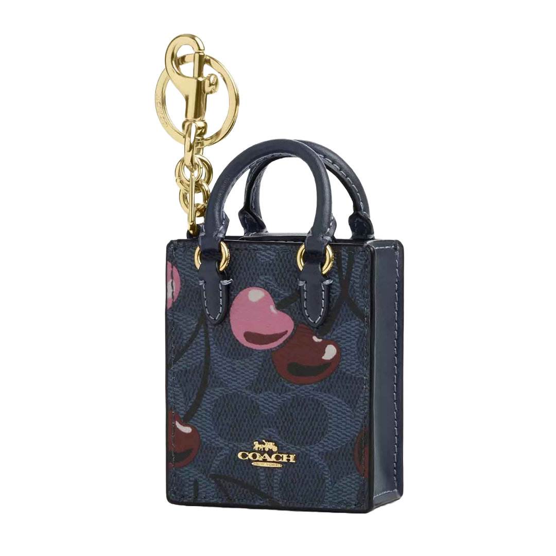 A Coach mini tote bag charm with a gold keychain, featuring a dark blue background decorated with colorful cherries and the brand's logo.