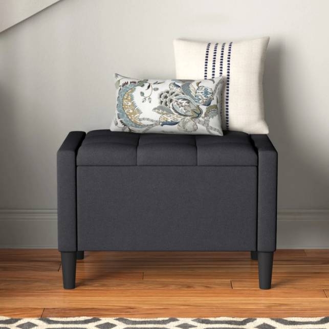 A dark gray upholstered storage bench with tufted detailing, supported by four wooden legs. It is topped with two decorative pillows, one featuring a floral pattern and the other with a striped design.
