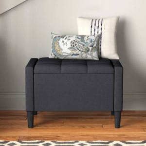 A dark gray upholstered storage bench with tufted detailing, supported by four wooden legs. It is topped with two decorative pillows, one featuring a floral pattern and the other with a striped design.