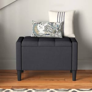 A dark gray upholstered storage bench with tufted detailing, supported by four wooden legs. It is topped with two decorative pillows, one featuring a floral pattern and the other with a striped design.
