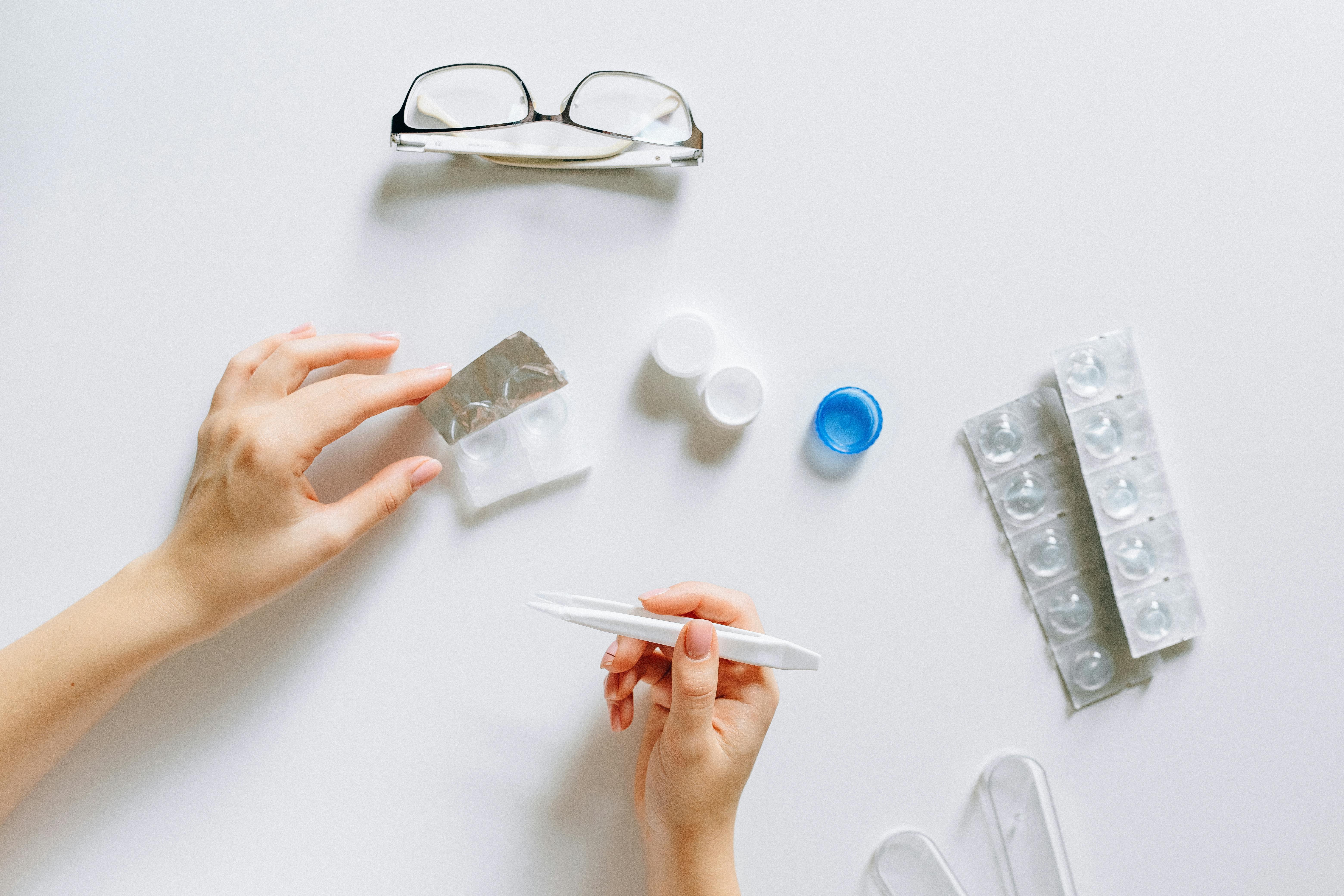 A person holds a pregnancy test with contact lens cases, glasses, and blister packs spread out on a white surface.