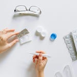 A person holds a pregnancy test with contact lens cases, glasses, and blister packs spread out on a white surface.