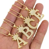 Gold necklaces with large, crown-topped letters \“A,\“ \“B,\“ and \“C\“ encrusted with small diamonds, displayed on a hand.