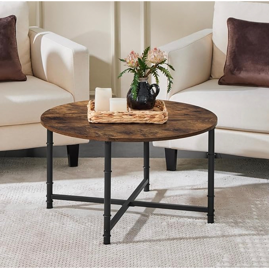 Round coffee table with a rustic wood top and black metal legs, featuring a woven tray holding candles and a black jug vase with flowers, placed between two cream-colored armchairs on a light textured rug.