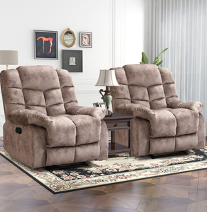 Two beige recliner chairs with plush cushioning are positioned on a floral rug. A wooden side table with a lamp and framed photos sits between them, against a white wall with artwork.