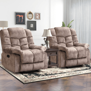 Two beige recliner chairs with plush cushioning are positioned on a floral rug. A wooden side table with a lamp and framed photos sits between them, against a white wall with artwork.