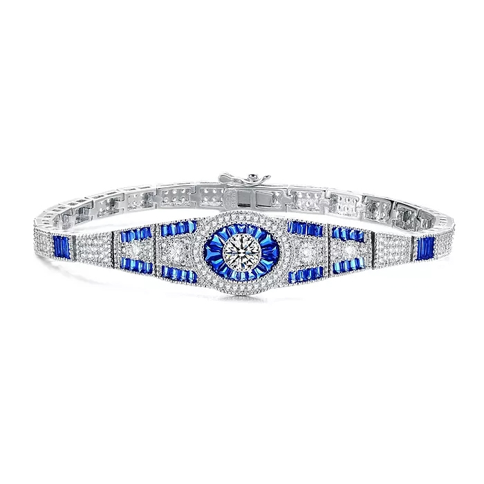 The Genevive Sterling Silver Blue CZ Bracelet features a central round clear cubic zirconia surrounded by blue baguette-cut stones and additional clear stones on a sterling silver band, creating an elegant, sparkling design.