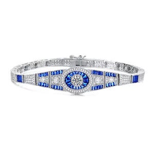 The Genevive Sterling Silver Blue CZ Bracelet features a central round clear cubic zirconia surrounded by blue baguette-cut stones and additional clear stones on a sterling silver band, creating an elegant, sparkling design.