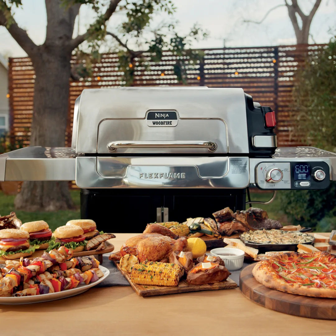 Ninja Woodfire FlexFlame Grill in stainless steel features a control panel with a digital display and knobs, surrounded by a spread of grilled food including burgers, chicken, corn, skewers, and pizza, set in an outdoor environment.