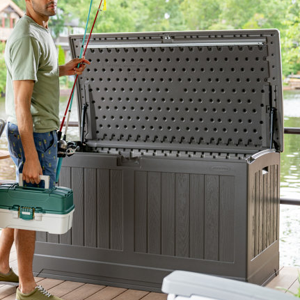 A dark gray Suncast deck box bench with a 200-gallon capacity, featuring a hinged lid with a pegboard pattern on the interior. It serves as storage, shown open with a person holding fishing gear nearby.