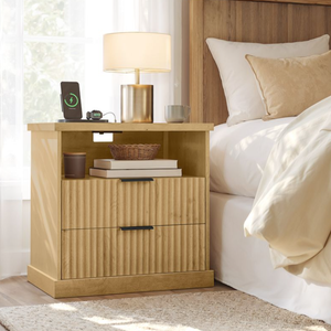 A wooden bedside table with a lamp, wireless phone charger, smart watch dock, mug, and basket. It features two ribbed drawers and an open shelf with books and a candle.