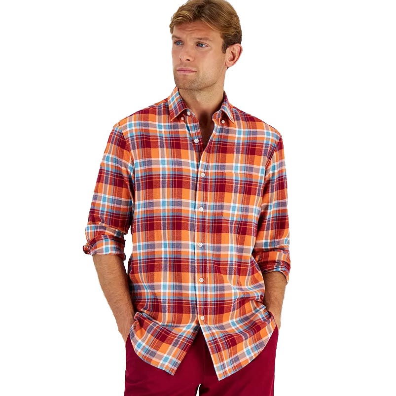 A man wears a Club Room regular-fit plaid flannel shirt, featuring a combination of red, orange, and blue checks. The shirt has a button-down front and long sleeves with the cuffs rolled up.