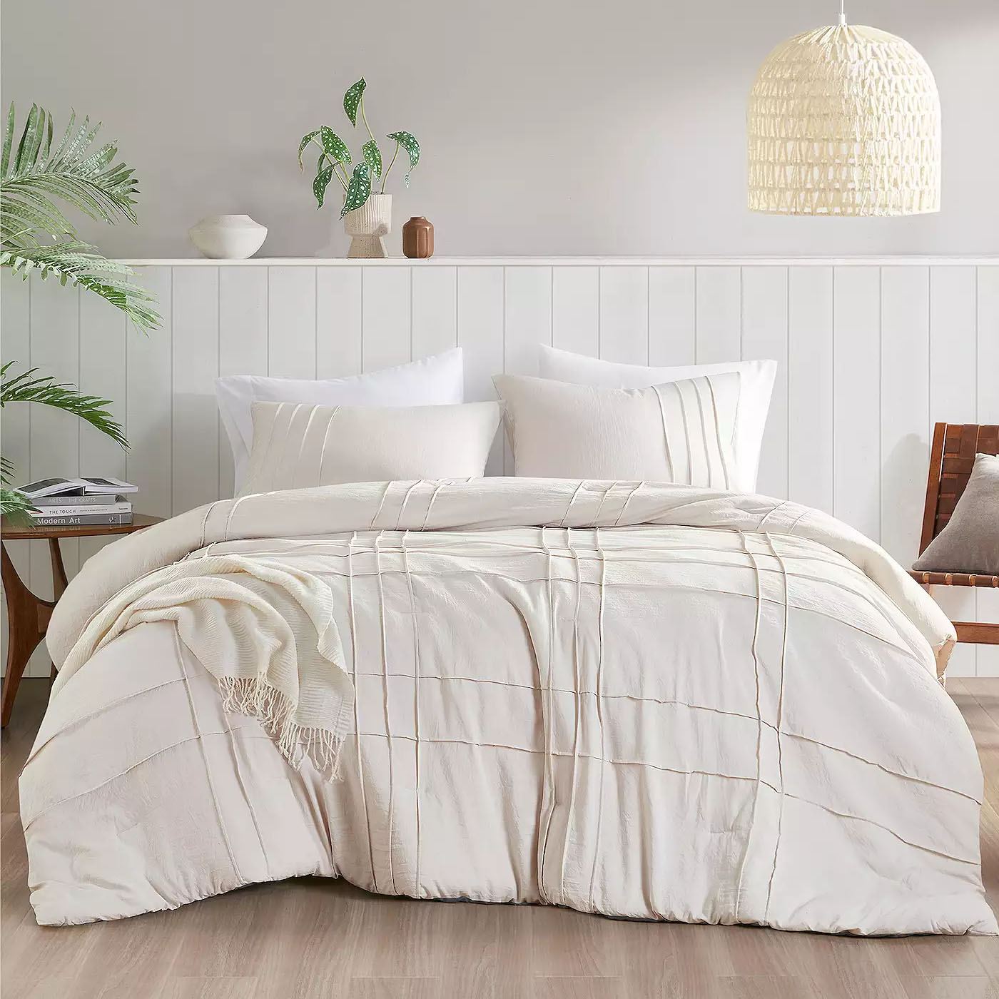 A cozy bedroom scene featuring a plush, textured duvet with matching pillows set against a white paneled headboard. The space is styled with a woven pendant light, green plants, and a wooden side chair for a minimalist, serene look.