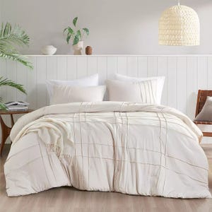A cozy bedroom scene featuring a plush, textured duvet with matching pillows set against a white paneled headboard. The space is styled with a woven pendant light, green plants, and a wooden side chair for a minimalist, serene look.