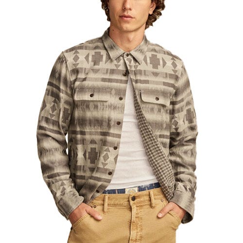 A man wears a Lucky Brand Jacquard Shirt featuring a geometric pattern in muted tones, with long sleeves and buttoned chest pockets, paired with tan pants and a white undershirt.