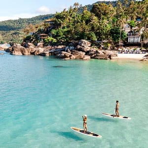 Two people are paddleboarding on clear, tropical waters near a beach with rocks and palm trees.