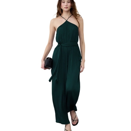 A woman wears a sleeveless dark green jumpsuit with a halter neck and wide-leg design, featuring a waist tie. She holds a black clutch and wears strappy black sandals.