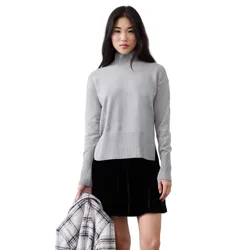 A woman is wearing a gray turtleneck sweater paired with a black velvet skirt. She holds a plaid coat, and the background is a textured, light-colored wall.
