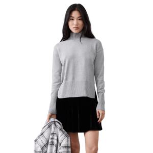 A woman is wearing a gray turtleneck sweater paired with a black velvet skirt. She holds a plaid coat, and the background is a textured, light-colored wall.
