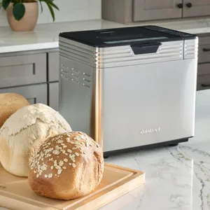 A stainless steel bread maker is on a kitchen counter next to three loaves of bread on a wooden board.